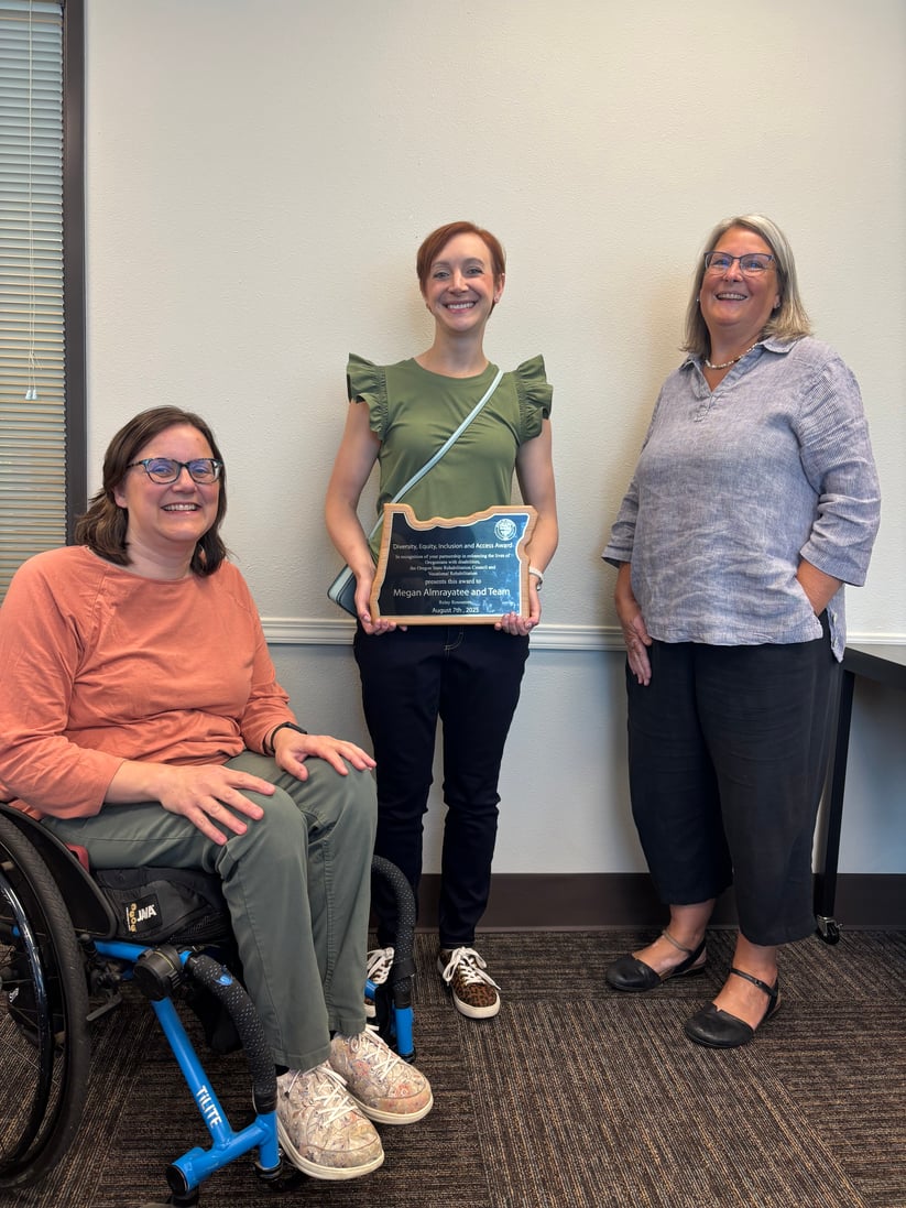 Megan holds an award in the shape of the state of Oregon as she receives the DEIA award from Susan and Erin 