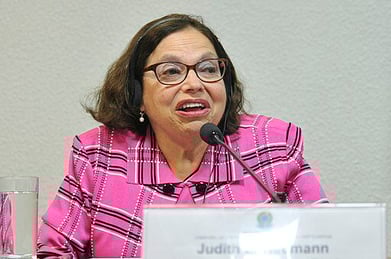 Judith Heumann wears a plaid pink shirt and speaks into a microphone