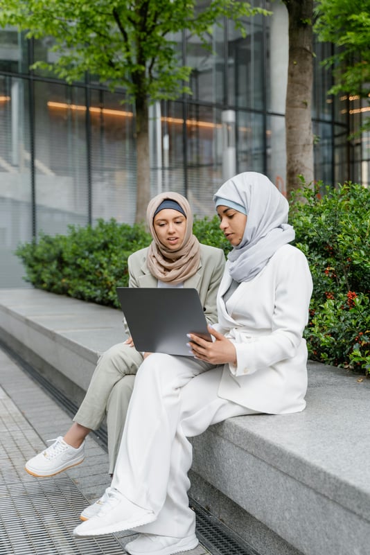 Two women in hijabs sit outside looking at a laptop.